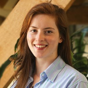 Portrait of a young woman with long auburn hair, smiling and wearing a light blue striped shirt.